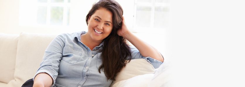 A smiling woman with long dark hair sits on a cream-coloured couch.