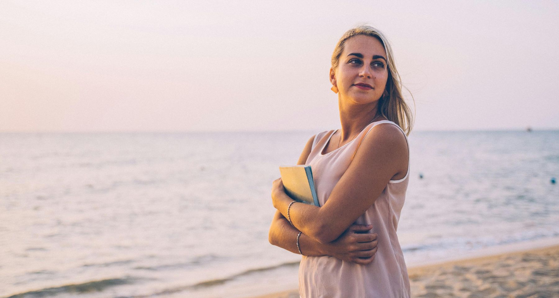 A blonde woman at the beach holding a notebook. 