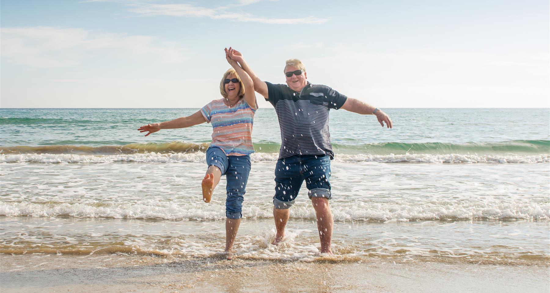 A happy older couple hold hands and kick up their heels in the shallows at a beach